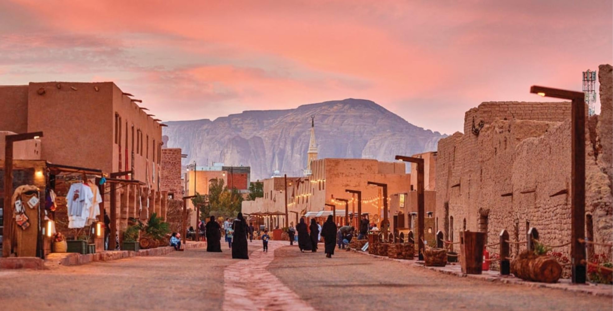 AlUla old town at sunset: low mud-brick buildings line a sandy lane against pink-orange sandstone cliffs, with figures walking through — the cultural landscape of the AlUla Cultural Heritage Impact Assessment.