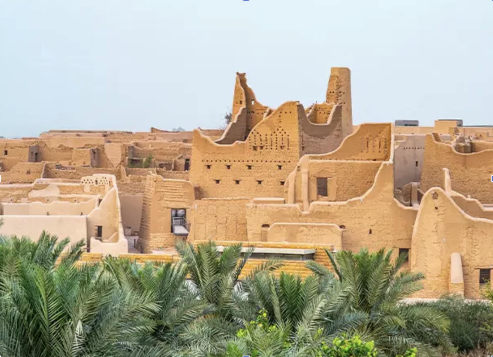 Ghasibah fortified district: ochre mud-brick towers and walls with crenellated parapets rise above a date-palm grove inside the UNESCO Al-Turaif buffer zone in Saudi Arabia.