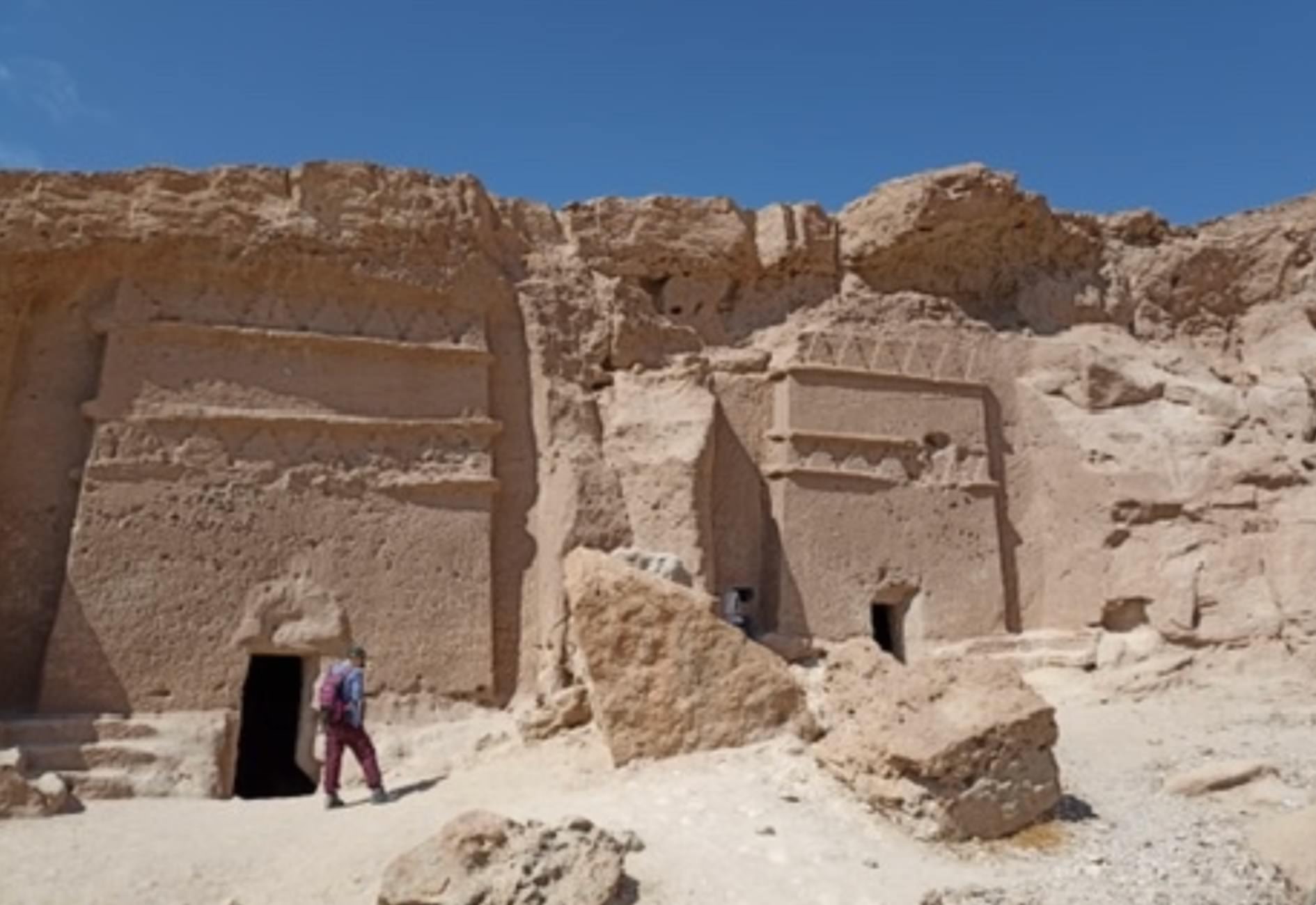 Two Nabataean rock-cut tomb facades carved into ochre sandstone cliffs at Mugha'ir Shu'ayb, NEOM, with a researcher walking past the entrance for scale.